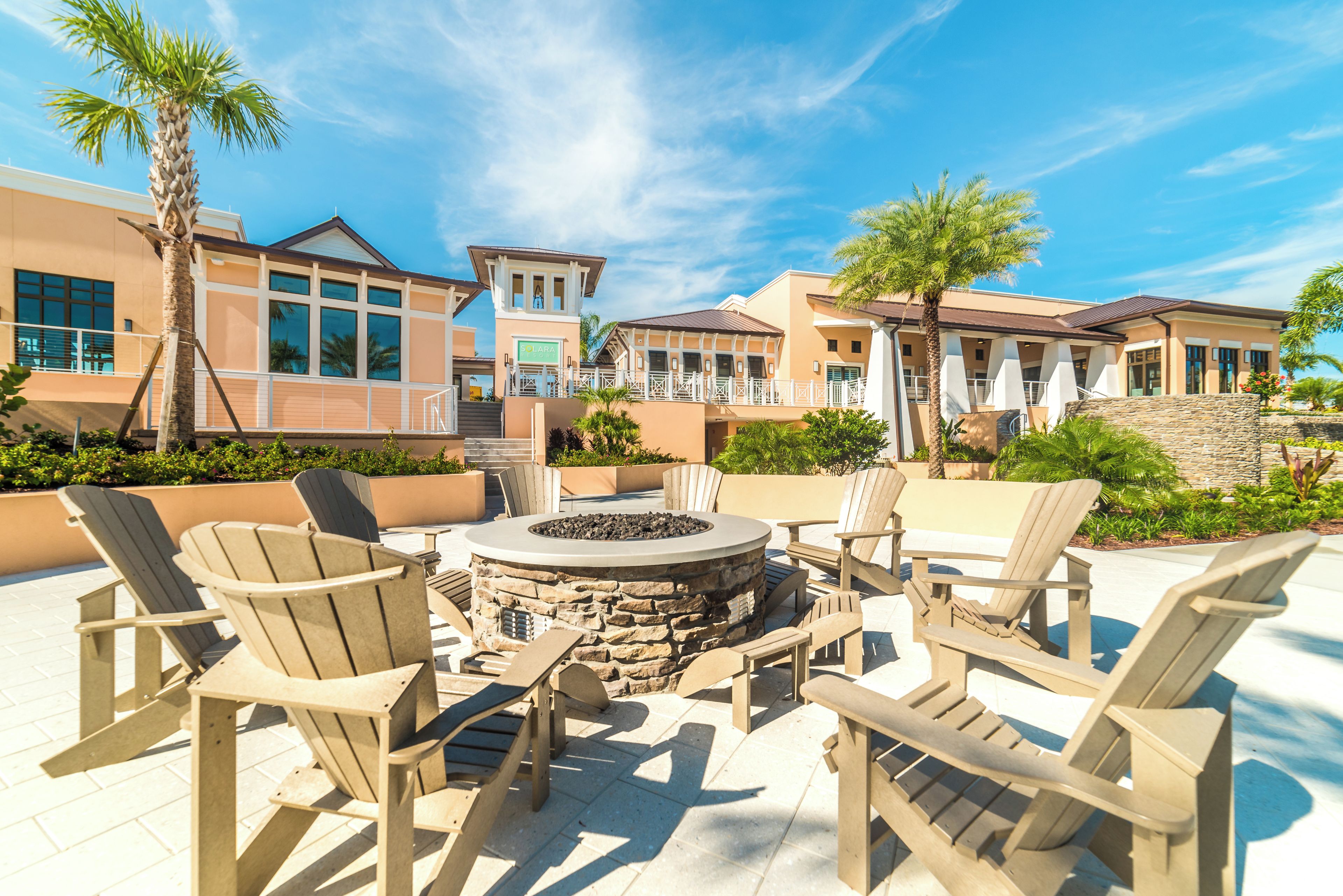 An outdoor seating area with Adirondack chairs and a stone fire pit, surrounded by palm trees and a building with large windows.