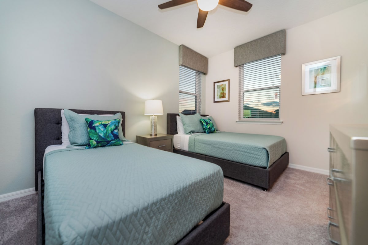 A bedroom with two beds, a nightstand, a lamp, a ceiling fan, and a window with blinds.