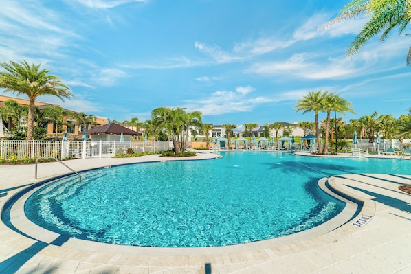 A clear blue pool surrounded by palm trees and lounge chairs, with a white fence and buildings in the background.