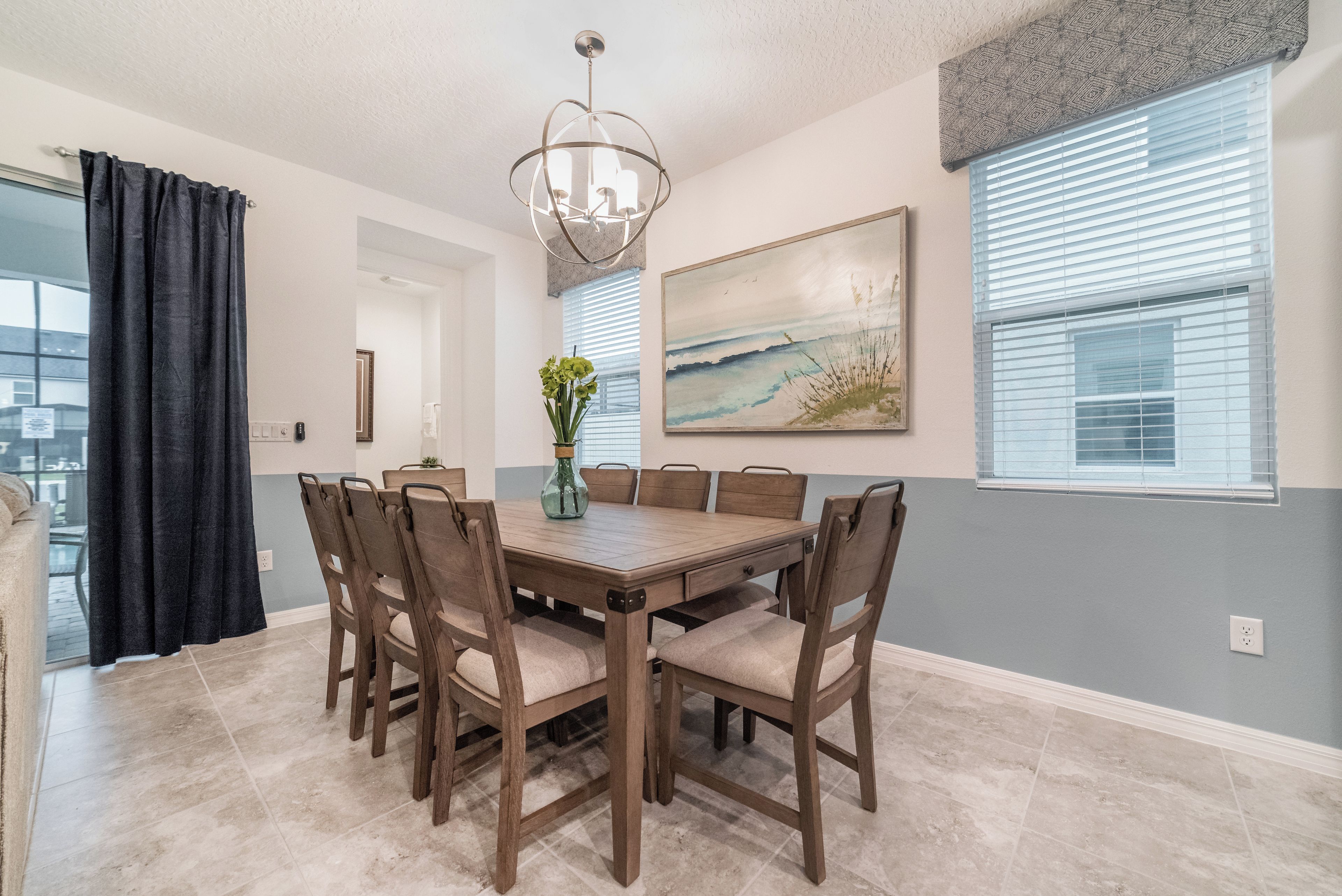 A dining area with a wooden table and chairs, a chandelier, a painting, and a window with blinds.