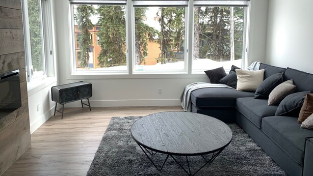 A modern living room with a grey sofa, a round coffee table, and a large window with a view of trees and buildings.