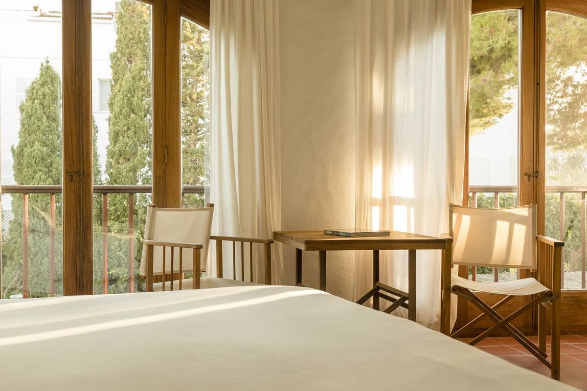 Un dormitorio con una cama grande, una mesita pequeña, dos sillas y un balcón con cortinas.