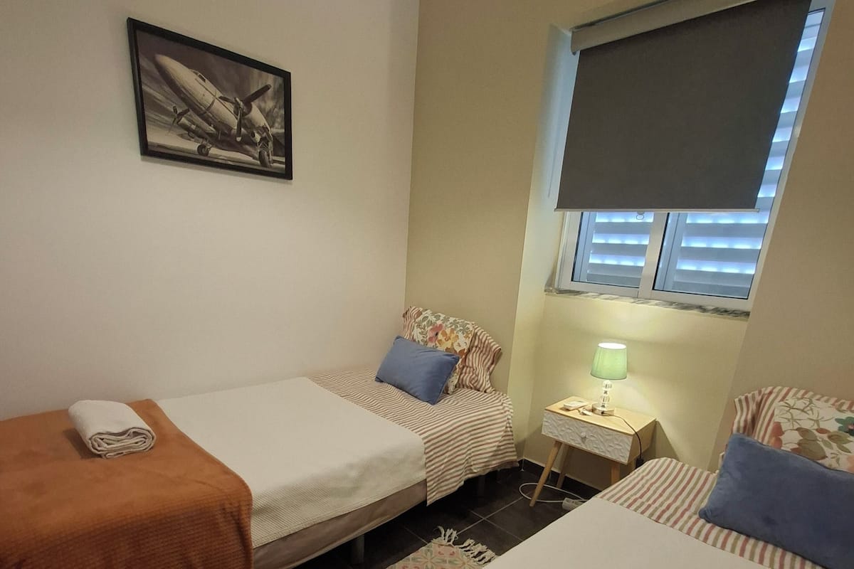 A hotel room with two beds, a nightstand with a lamp, a window with blinds, and a framed picture on the wall.
