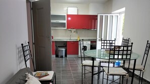A modern living room with a glass dining table and chairs, a grey sofa with a colorful cushion, and a kitchen in the background with red cabinets. - Sleep & Go Faro Airport Guest House (Faro)