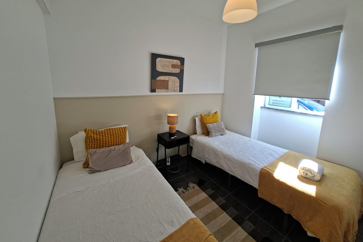 A hotel room with two single beds, a small table with a lamp, a window with a view, and a ceiling light.