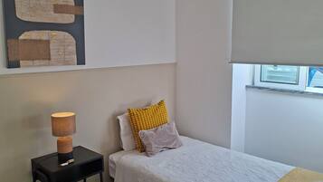 Een kleine, verzorgde slaapkamer met een enkel bed, een nachtkastje met een lamp, een klein tafeltje met een handdoek, en een raam met een luxaflex.