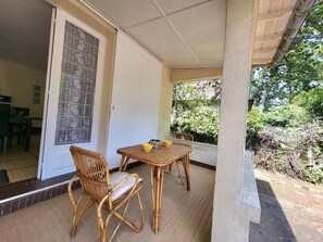 A covered patio area with a table and two wicker chairs, a door with a decorative glass panel, and a view of greenery outside.