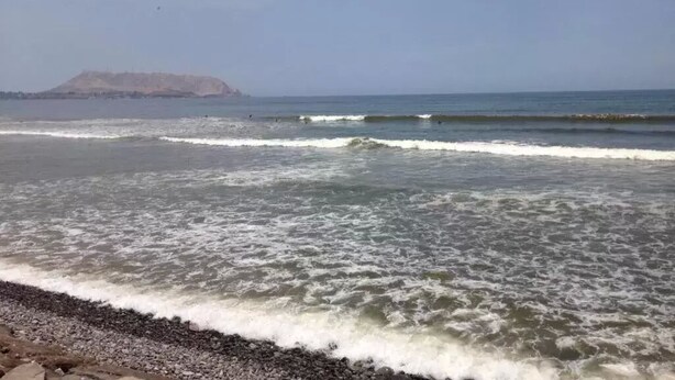 Una playa con olas, piedras y una isla a lo lejos.