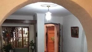 Sala de estar con un sofá, una mesa de centro y una mesita de noche con lámpara. Se ve una puerta que da a otra habitación y una ventana con cortinas.