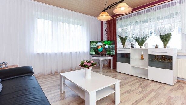 Una sala moderna con un sofá negro, una mesa de centro blanca con un florero y un televisor sobre un mueble, además de una zona de cocina con gabinetes blancos y elementos decorativos.