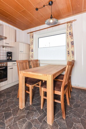 Una mesa de comedor de madera con sillas, una cocina con armarios de madera clara, suelo de baldosas de piedra y una ventana con cortinas.