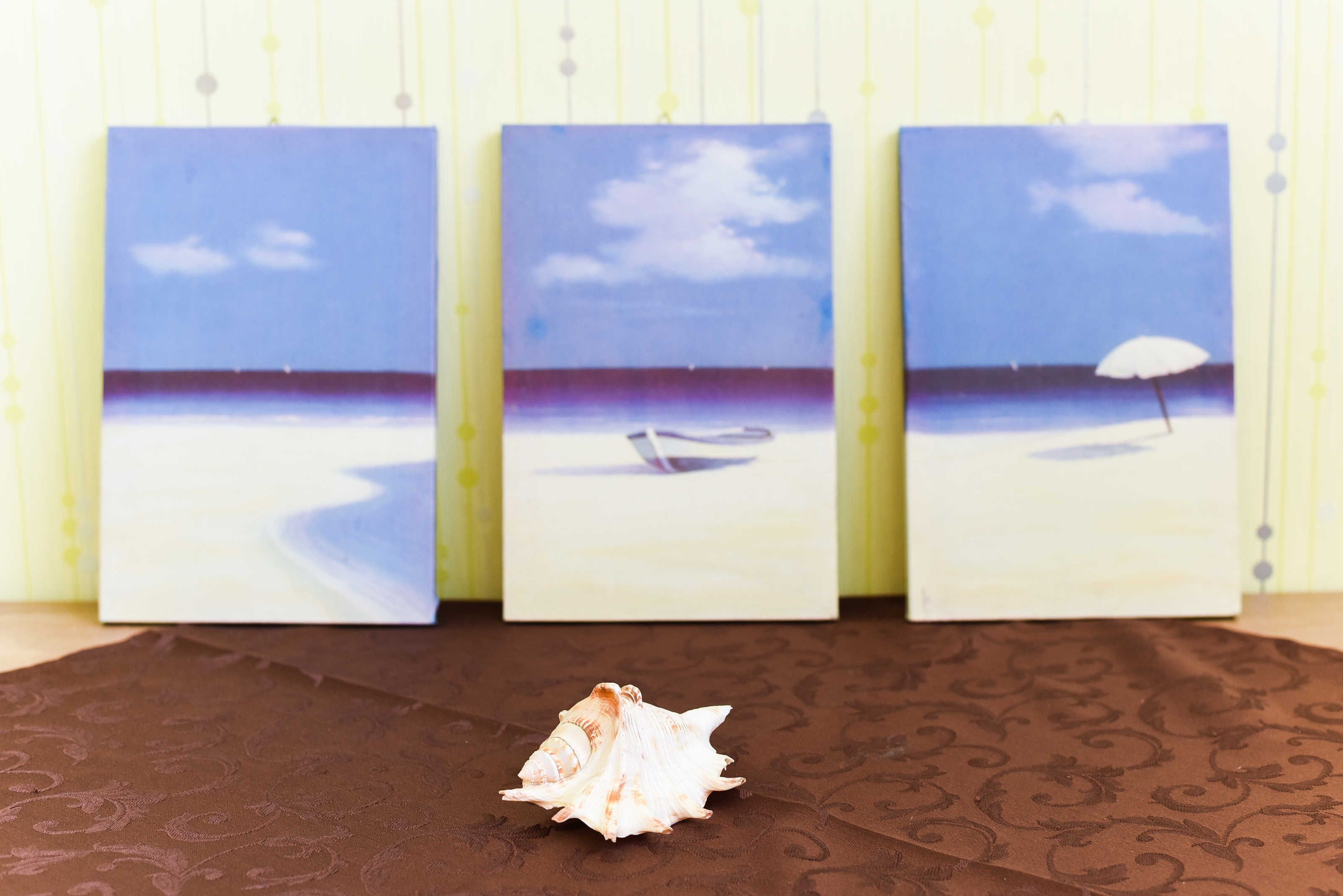 Three framed paintings of a beach scene with a seashell in front.