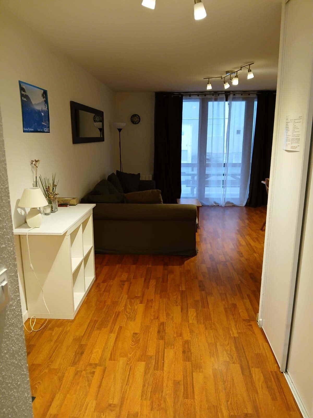 Un salon avec un canapé, une petite table blanche surmontée d’une lampe, et une grande fenêtre avec des rideaux.