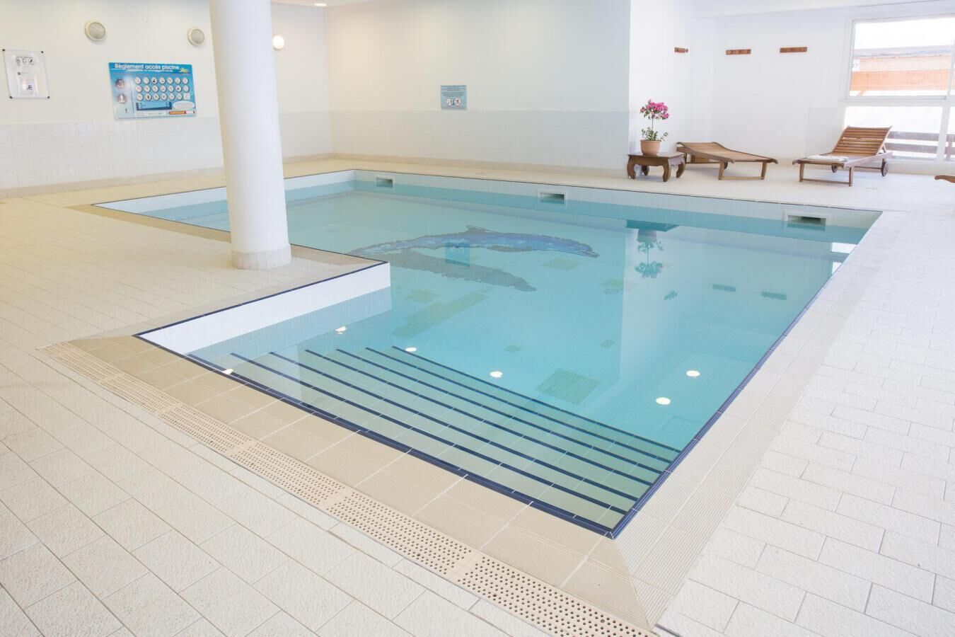 An indoor swimming pool with a tiled floor, a pool ladder, and lounge chairs.