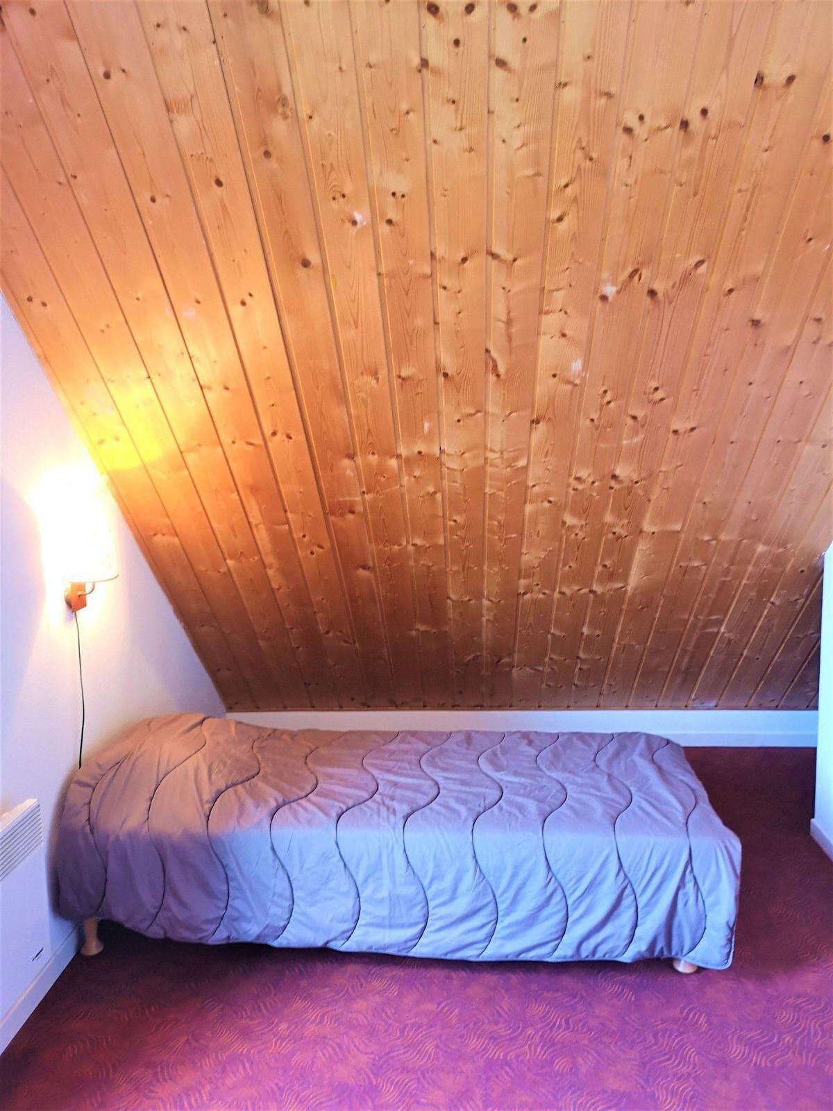 Ein kleiner Raum mit schräg verlaufender Holzbalkendecke, einem Einzelbett mit gemustertem Überwurf und einer wandmontierten Leuchte.