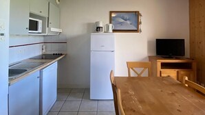 Cozinha compacta com micro-ondas, geladeira e cafeteira. Há uma mesa de jantar de madeira com cadeiras e uma televisão sobre um móvel de madeira.