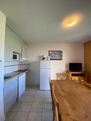 Une cuisine compacte équipée d’un four à micro-ondes, d’un réfrigérateur et d’une machine à café. On y trouve une table à manger en bois avec des chaises, ainsi qu’un téléviseur posé sur un meuble en bois.