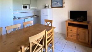 Uma cozinha compacta com mesa e cadeiras, um frigorífico pequeno, um micro-ondas e uma televisão sobre um móvel de madeira.