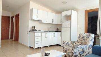 Una cocina moderna con gabinetes blancos, una estufa empotrada y un refrigerador. Hay un sillón con estampado floral y una mesa de centro blanca con un letrero encima.