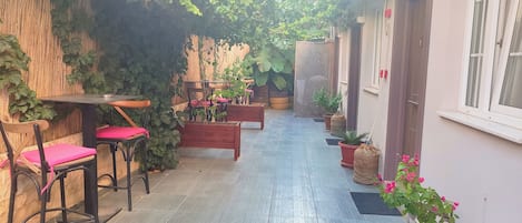 A narrow outdoor corridor with wooden benches and tables, potted plants, and a tiled floor.
