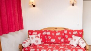 Um sofá-cama de madeira com capa estampada em vermelho e branco, duas luminárias fixadas na parede e uma mesinha com copos e uma vela.