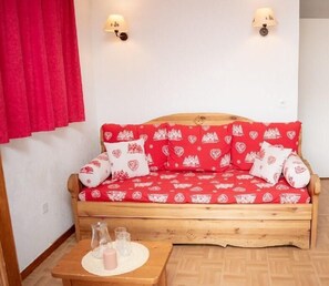 A wooden sofa bed with a red and white patterned cover, two wall-mounted lamps, and a small table with glasses and a candle.