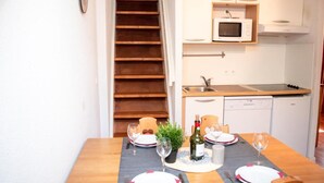 A dining area with a wooden table set for two, a small potted plant, and a bottle of wine. A kitchen with a microwave and dishwasher is visible in the background. - Wifi, 1st floor, swimming pool, balcony, tv, 45m², Saint Lary Soulan (Vignec)