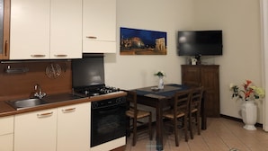 Une cuisine compacte avec des meubles blancs, une cuisinière et un évier. On y trouve une table à manger avec des chaises et un téléviseur à écran plat fixé au mur.