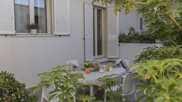 Un piccolo patio con un tavolo apparecchiato per due, circondato da piante in vaso e un recinto bianco.