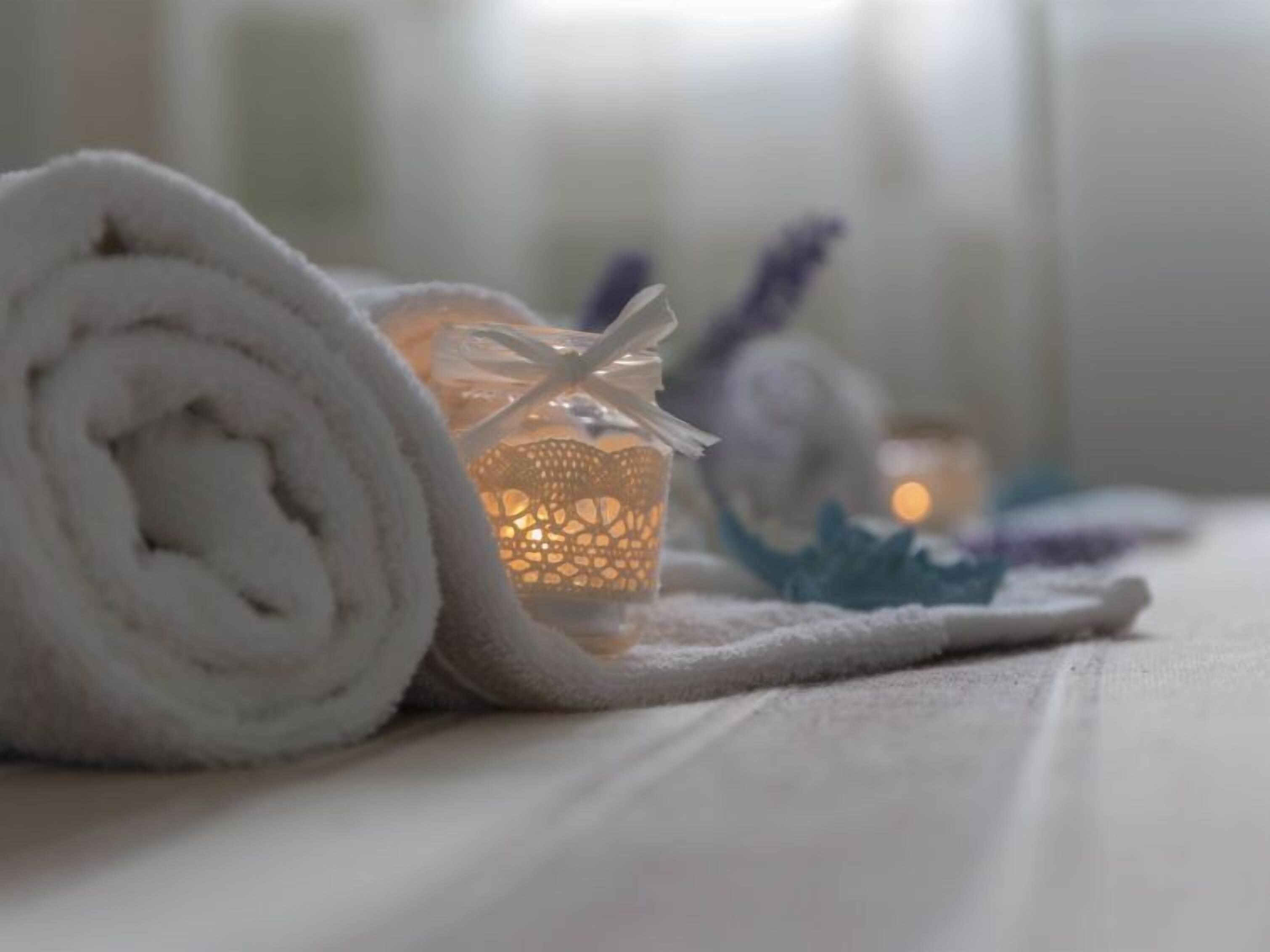 A neatly rolled white towel on a bed with a lit candle and a decorative item in the background.