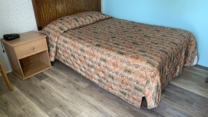 A bedroom with a wooden headboard, a bed with a patterned bedspread, a wooden nightstand with a drawer, and a wooden floor. - Erwin Motel (Painted Post)