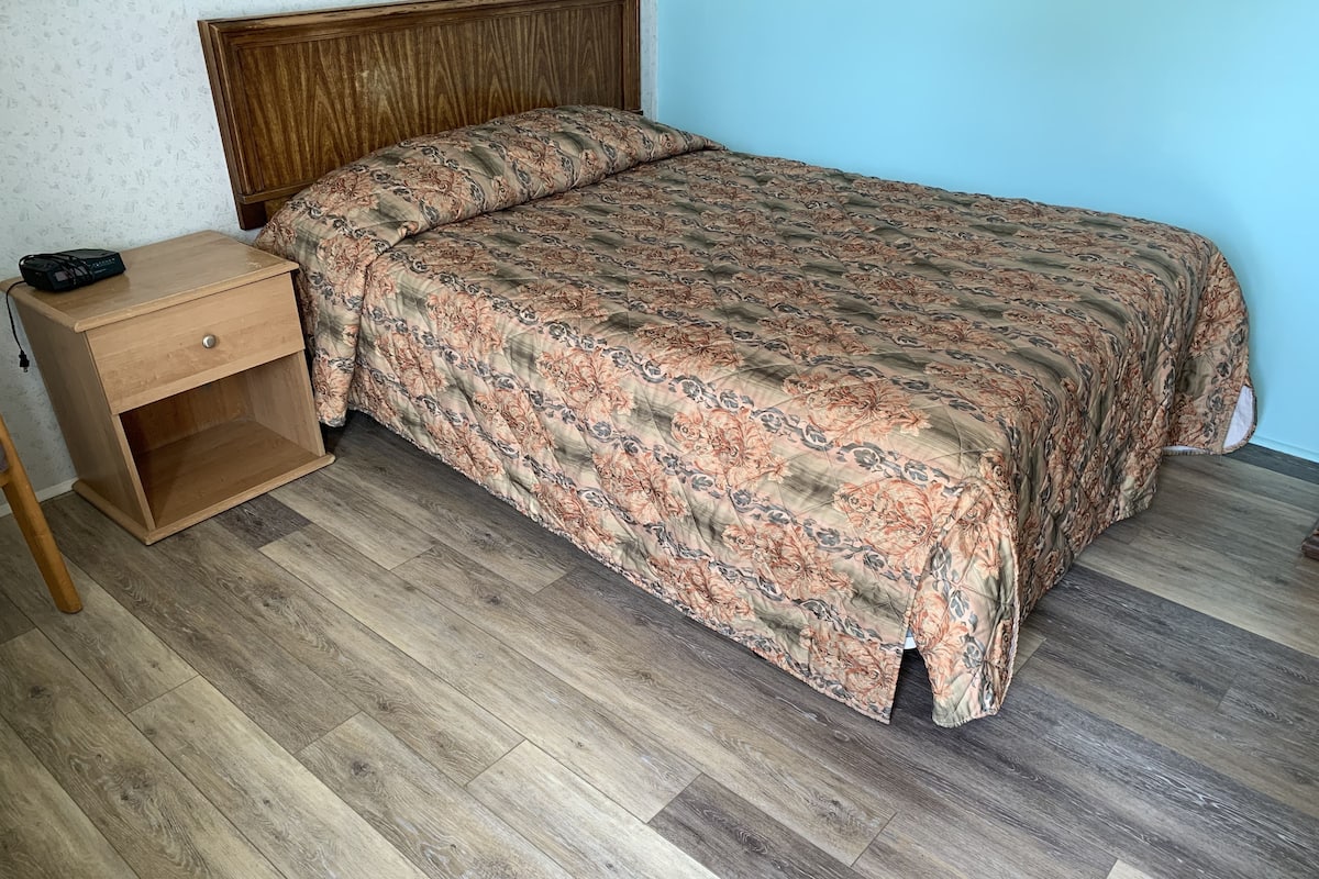 A bedroom with a wooden headboard, a bed with a patterned bedspread, a wooden nightstand with a drawer, and a wooden floor.