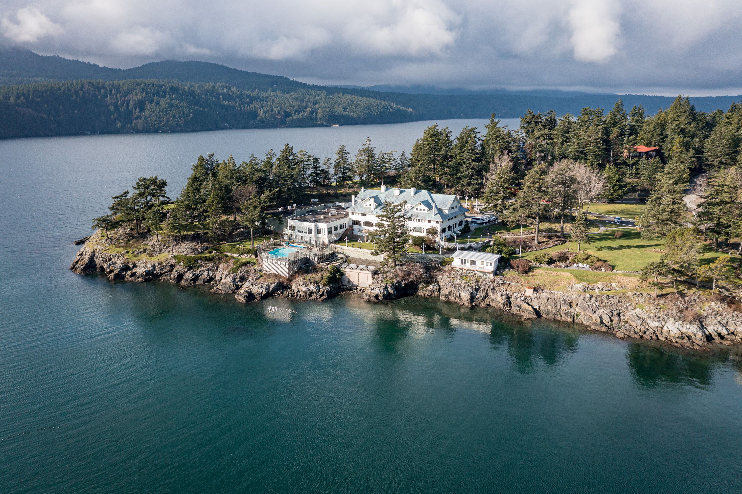 A large, multi-story building with a prominent roof and multiple windows, situated on a rocky shoreline with a body of water in the background.
