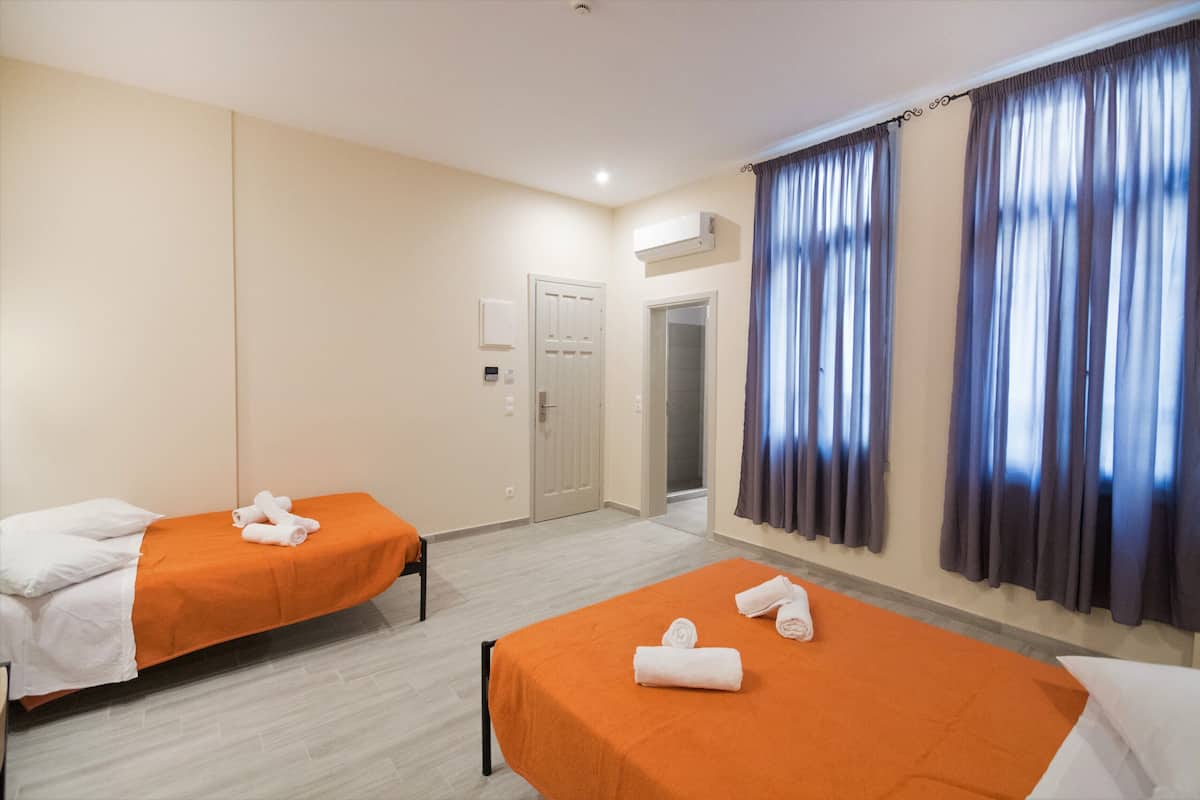 A hotel room with two single beds, each with an orange bedspread and white pillows. There is a door leading to a bathroom, and a window with dark curtains.