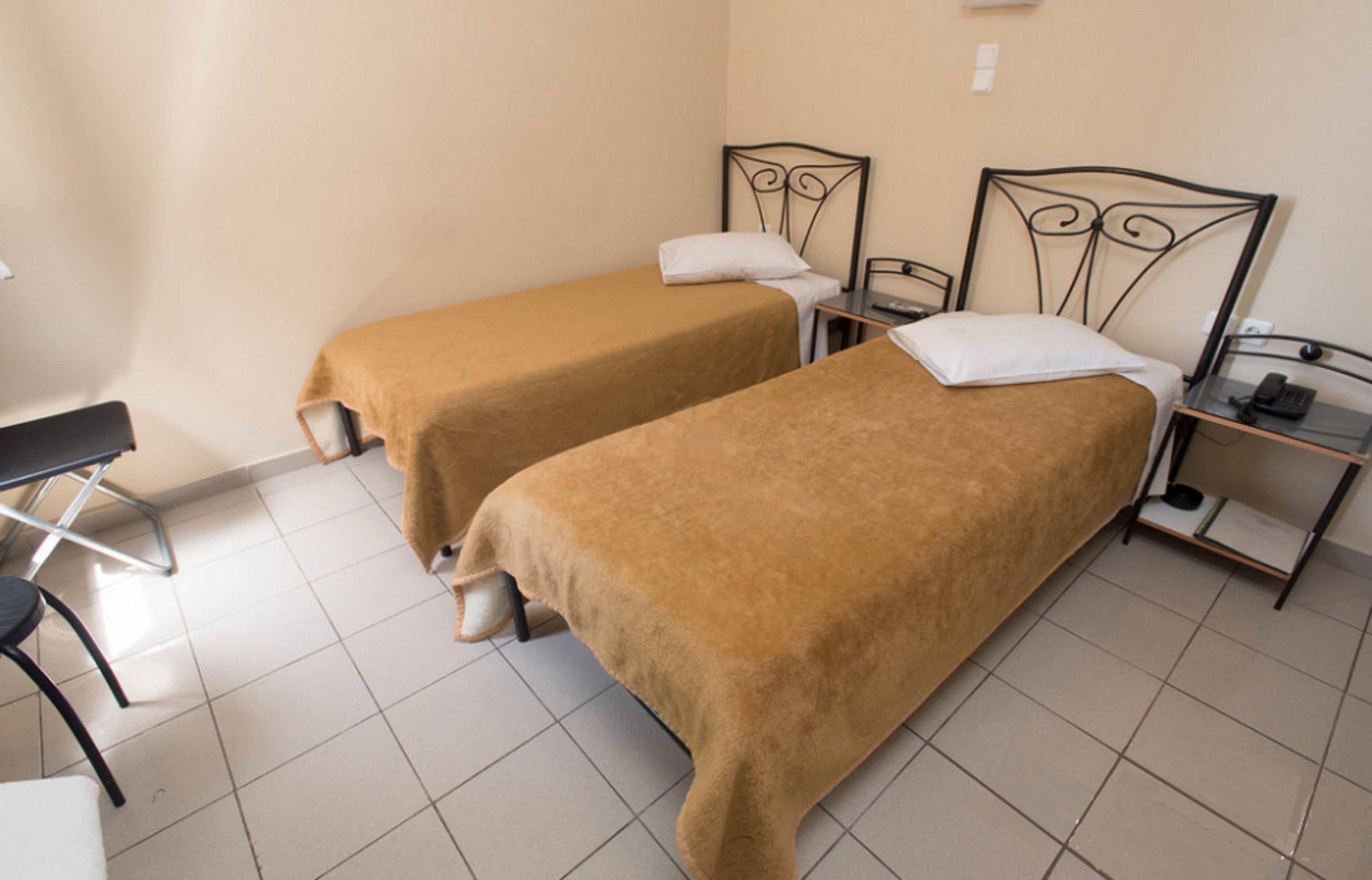 Deux lits simples avec des cadres en métal, une petite table et une chaise dans une pièce dont le sol est recouvert de carreaux.