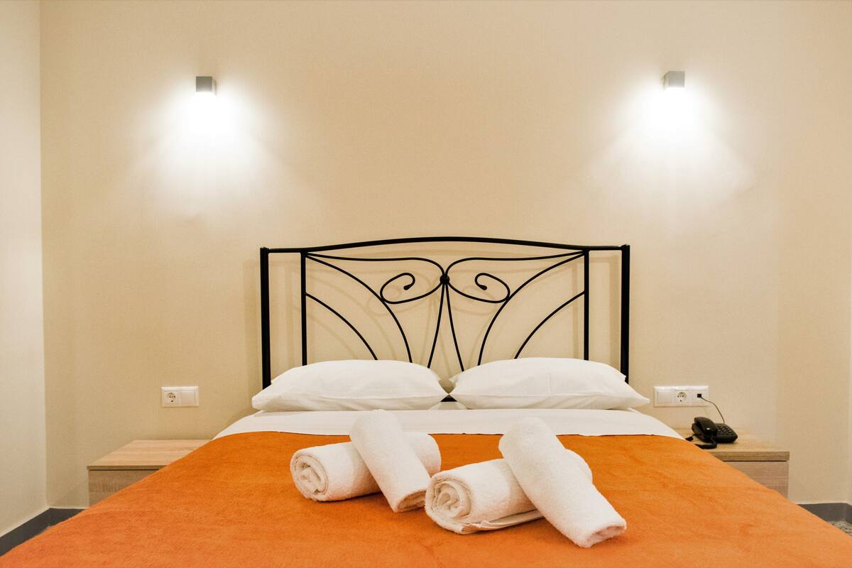 A neatly made bed with an orange bedspread and white pillows, two rolled towels on the bed, and a nightstand with a phone.