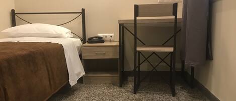 A bedroom with a bed, a desk with a chair, a nightstand, and a power outlet on the wall.
