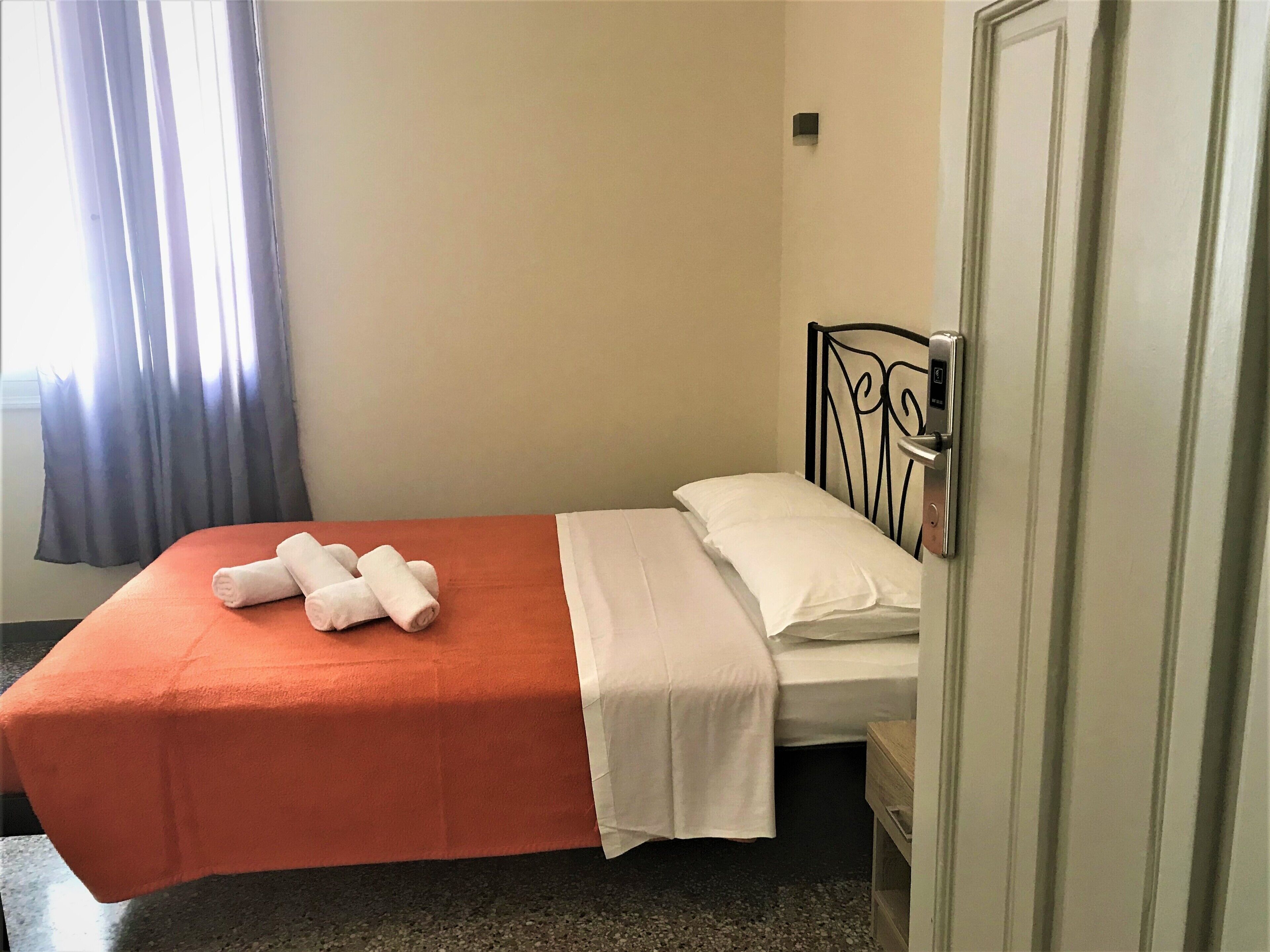 Una cama individual con colcha naranja y ropa de cama blanca, una mesita de noche pequeña y una puerta con picaporte.