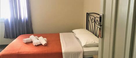 Una cama individual con colcha naranja y ropa de cama blanca, una mesita de noche pequeña y una puerta con picaporte.