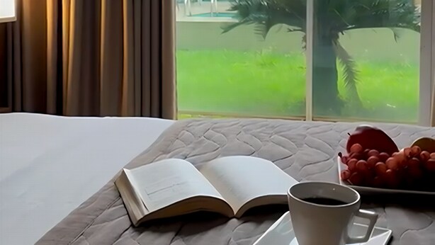 A hotel room with a bed, an open book, a cup of coffee, and a view of a building and greenery outside. - Oasis Hotel (Araguaina)