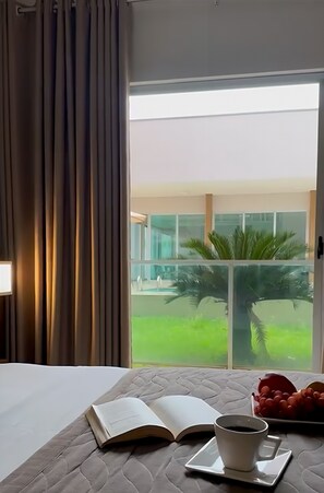 A hotel room with a bed, an open book, a cup of coffee, and a view of a building and greenery outside. - Oasis Hotel (Araguaina)