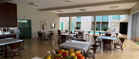 A dining area with a fruit bowl on a pedestal, tables, chairs, and a view of the outside.