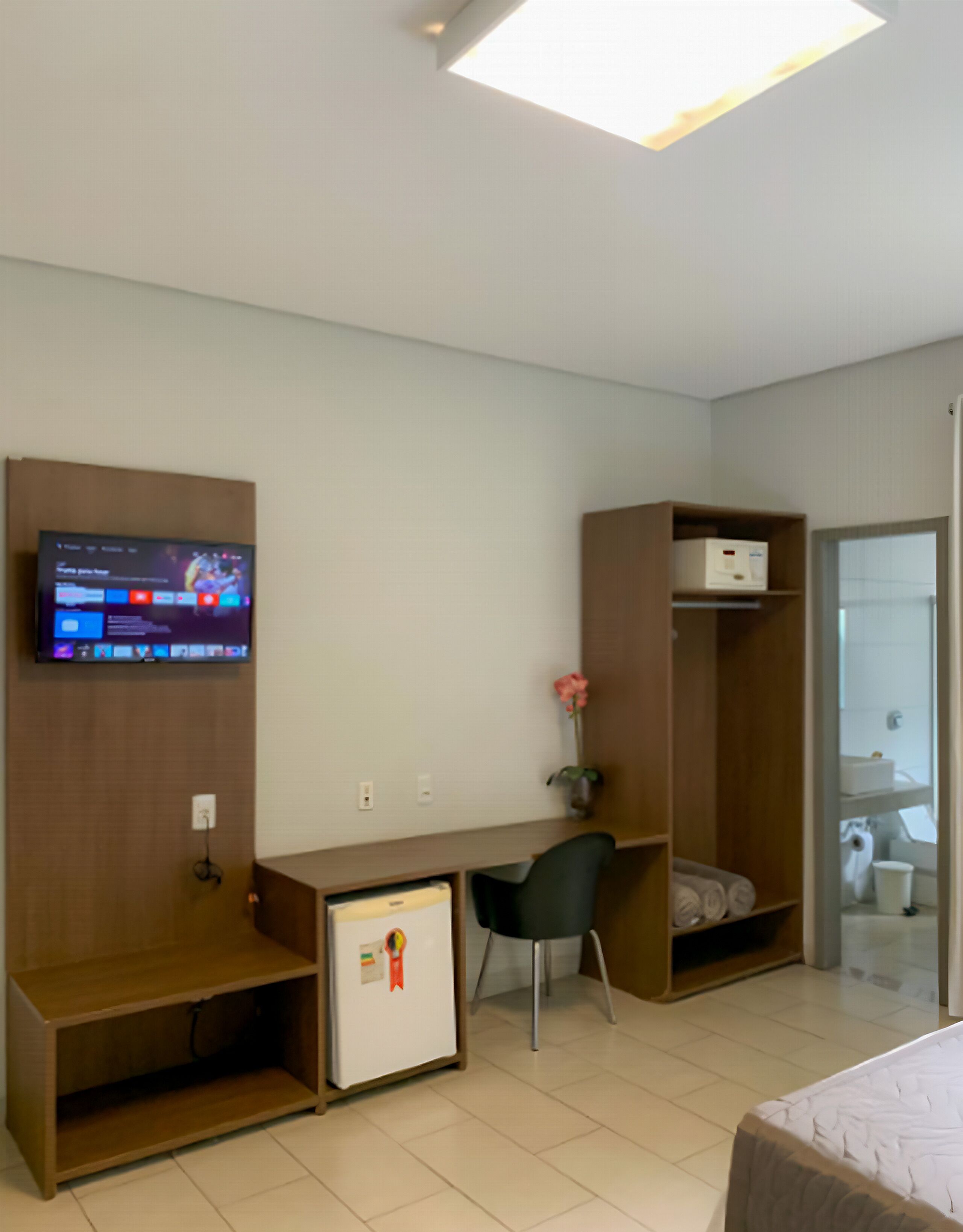 A hotel room with a bed, a desk with a computer, a TV, a small fridge, and a wardrobe.