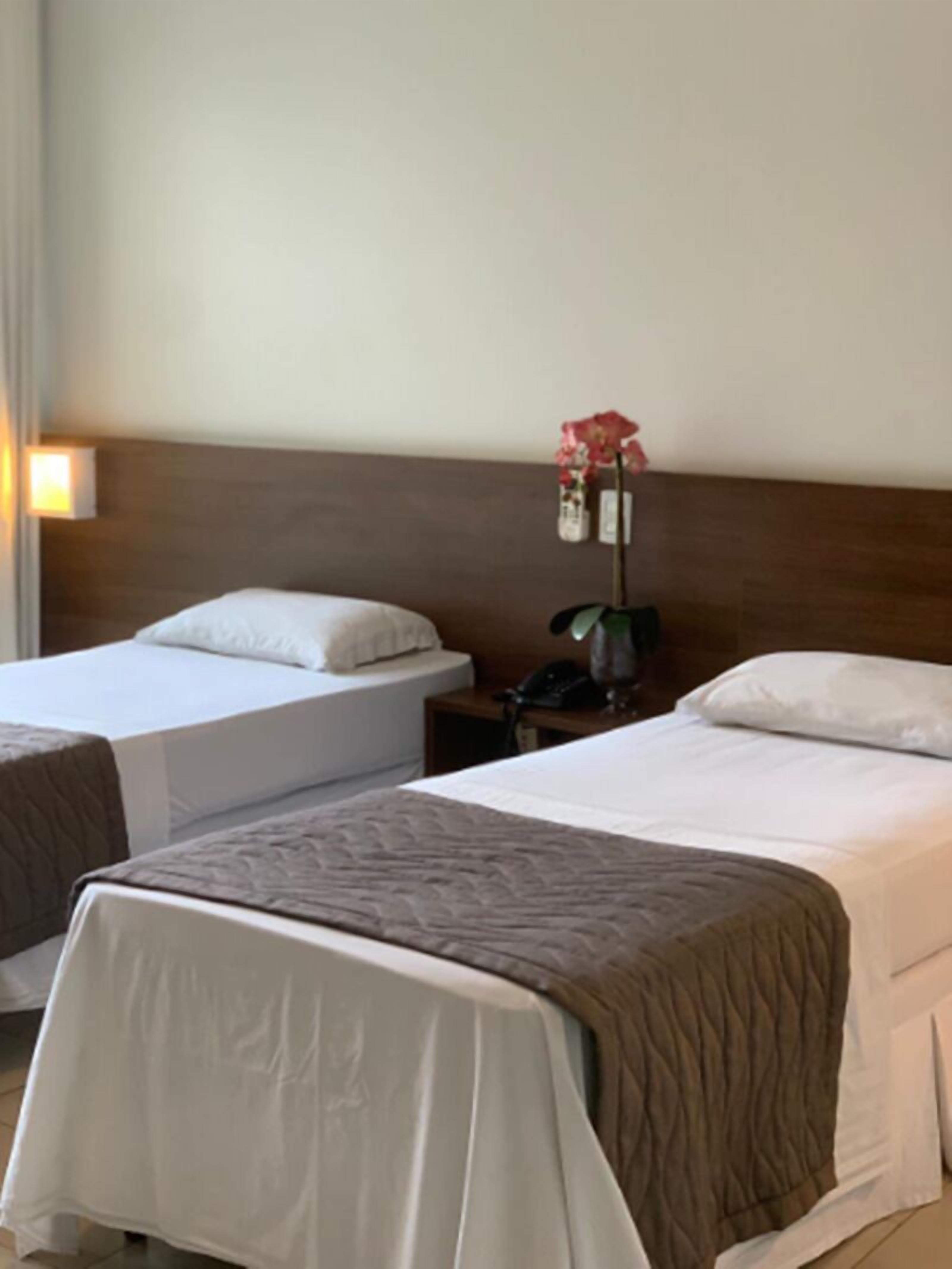 Habitación de hotel con dos camas, una mesita de noche con teléfono, una lámpara de pared y un florero con flores.