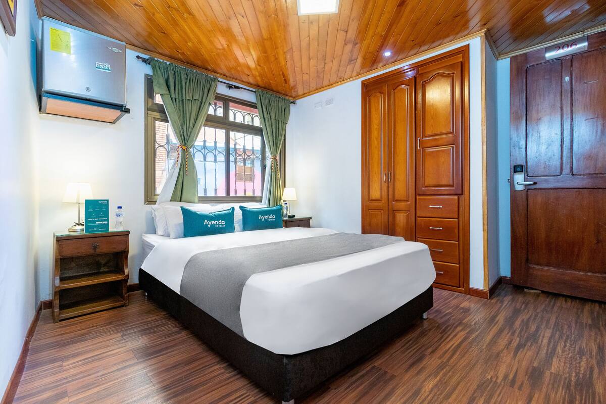 A hotel room with a wooden ceiling, a bed with white and grey bedding, a nightstand with a lamp, and a wardrobe.