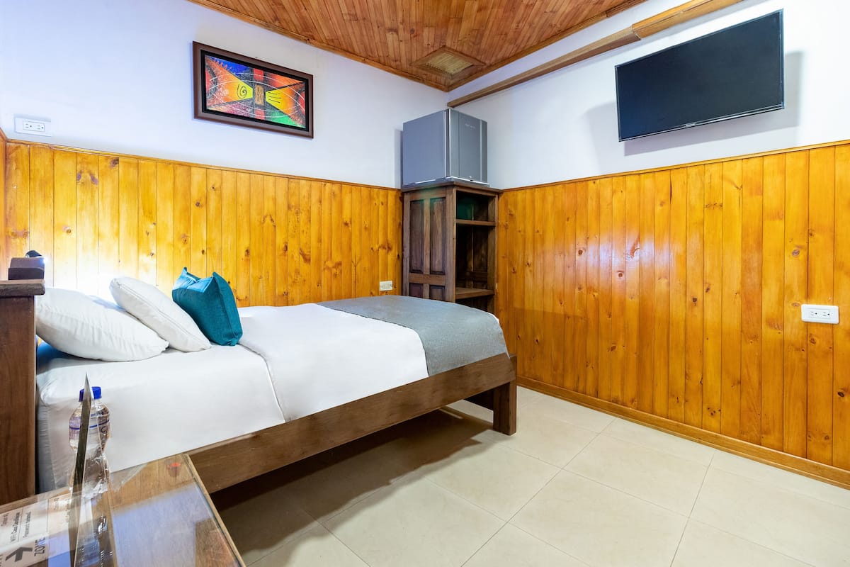 A bedroom with wooden paneling, a bed with white and teal bedding, a TV mounted on the wall, and a small shelf unit.
