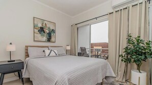 Een hotelkamer met een groot bed, een nachtkastje, een lamp, een schilderij, een potplant en uitzicht op buiten.