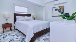 Een slaapkamer met een bed, twee nachtkastjes, een raam, een plant en een aan de muur bevestigde airconditioner.