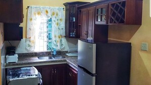A compact kitchen with dark wooden cabinets, a stainless steel refrigerator, a built-in oven, and a window with a floral curtain. - I Heart JA Me Lo Dio Dios Ocho Rios (Ocho Rios)