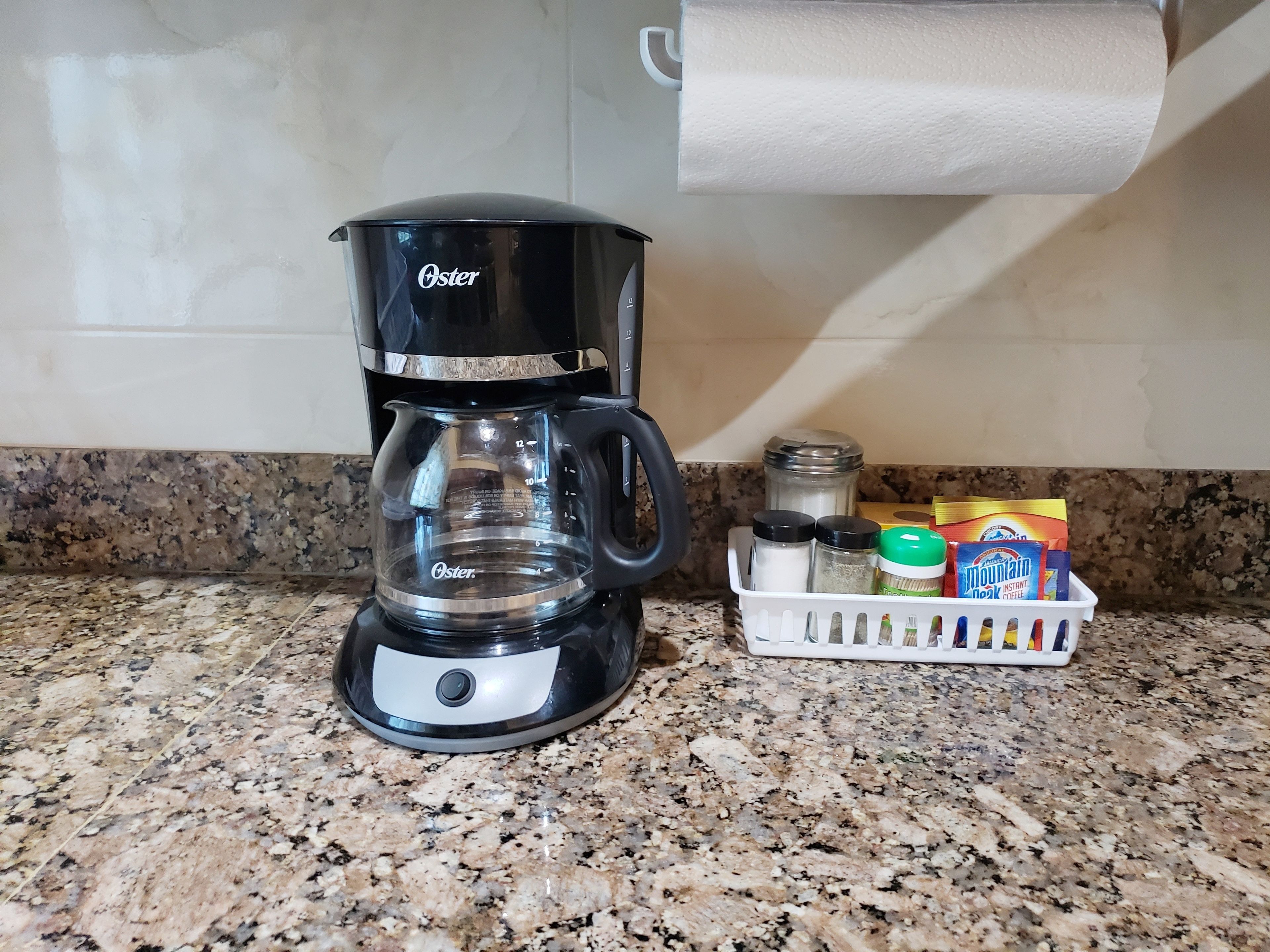 A black Oster coffee maker on a granite countertop with a basket of coffee pods and spices nearby.